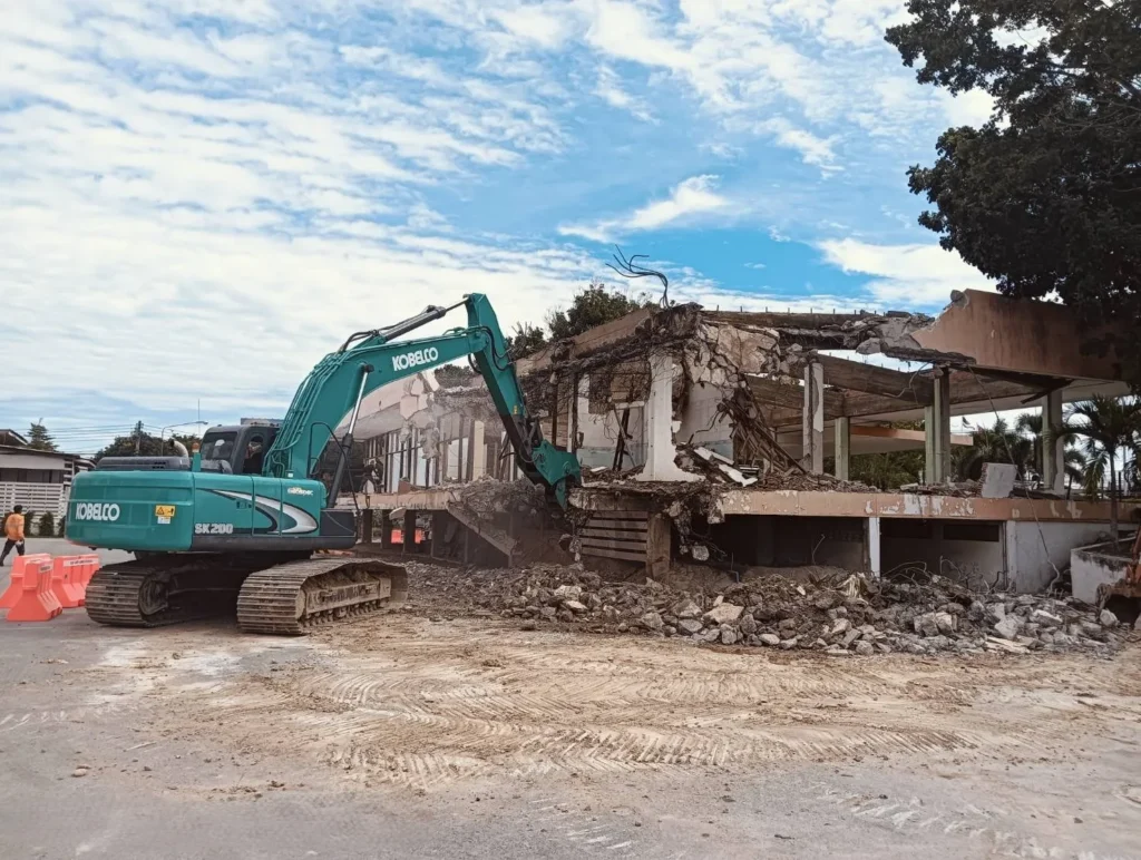 Teal Kobelco excavator tearing down a concrete building, rubble on the ground, construction barriers nearby under a cloudy sky